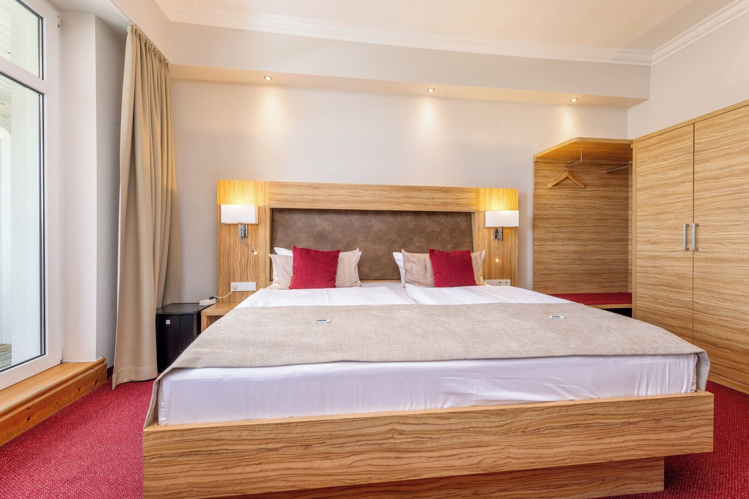A hotel room with a large bed, wooden headboard, two bedside lamps, a wardrobe, and a window with curtains.