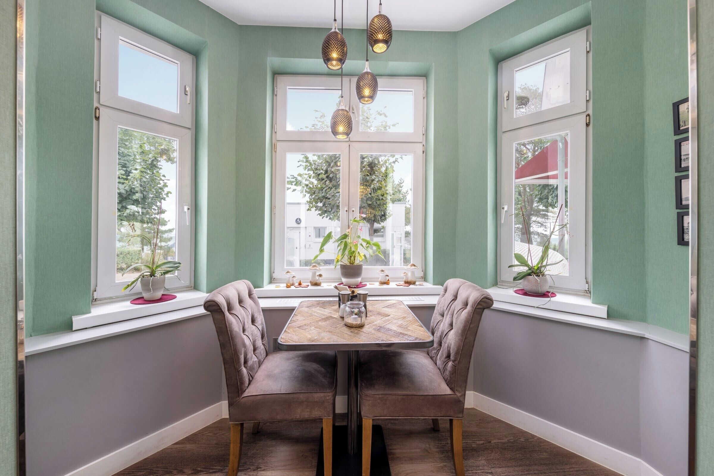 A dining area with a table and chairs, large windows with plants, and hanging pendant lights.