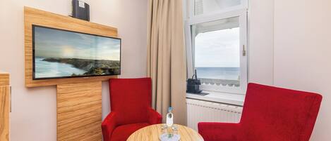 A hotel room with a wooden TV stand, a painting, two red chairs, a small round table with a bottle, and a window with a view of the sea.