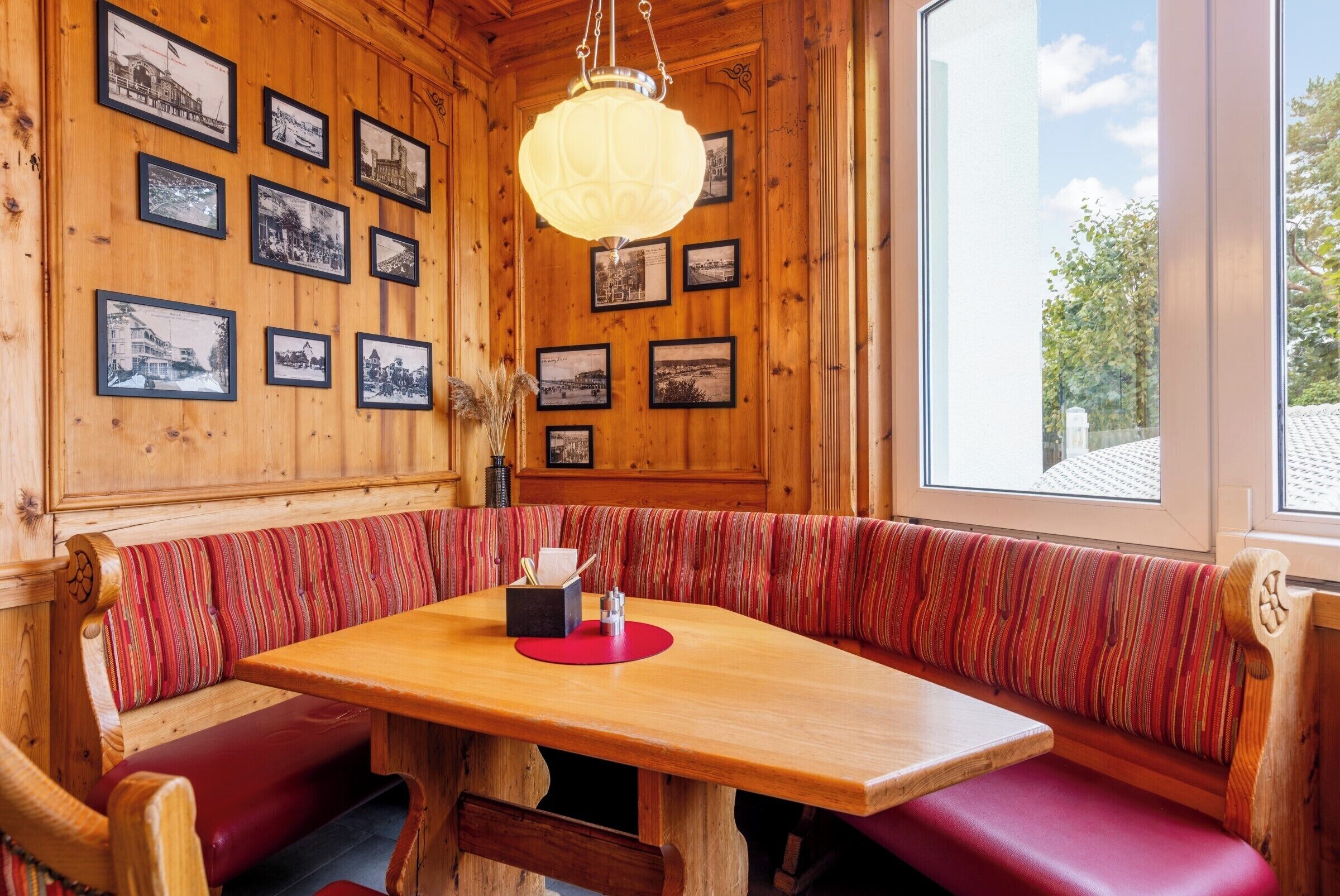 Un interior de madera con una zona de estar con banco, una mesa con camino de mesa rojo y fotografías en blanco y negro enmarcadas en las paredes.