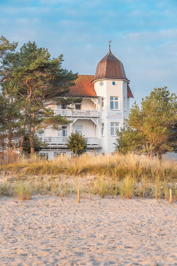 A white building with a brown dome roof and a balcony, surrounded by sand and sparse vegetation. - Strandhotel zur Promenade (Binz)