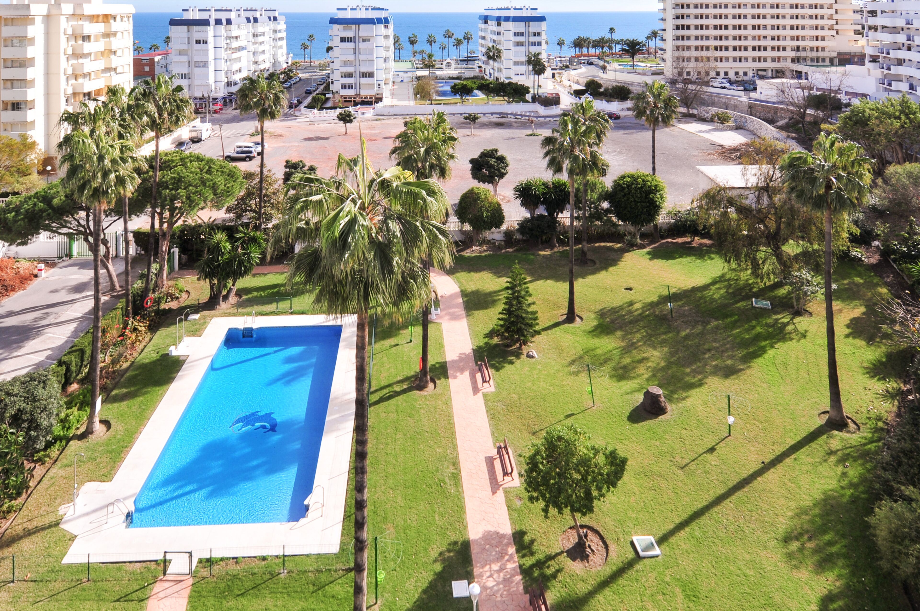 Une piscine entourée d’une pelouse bien entretenue et agrémentée de palmiers, le tout sur fond d’immeubles de grande hauteur et d’un ciel dégagé.