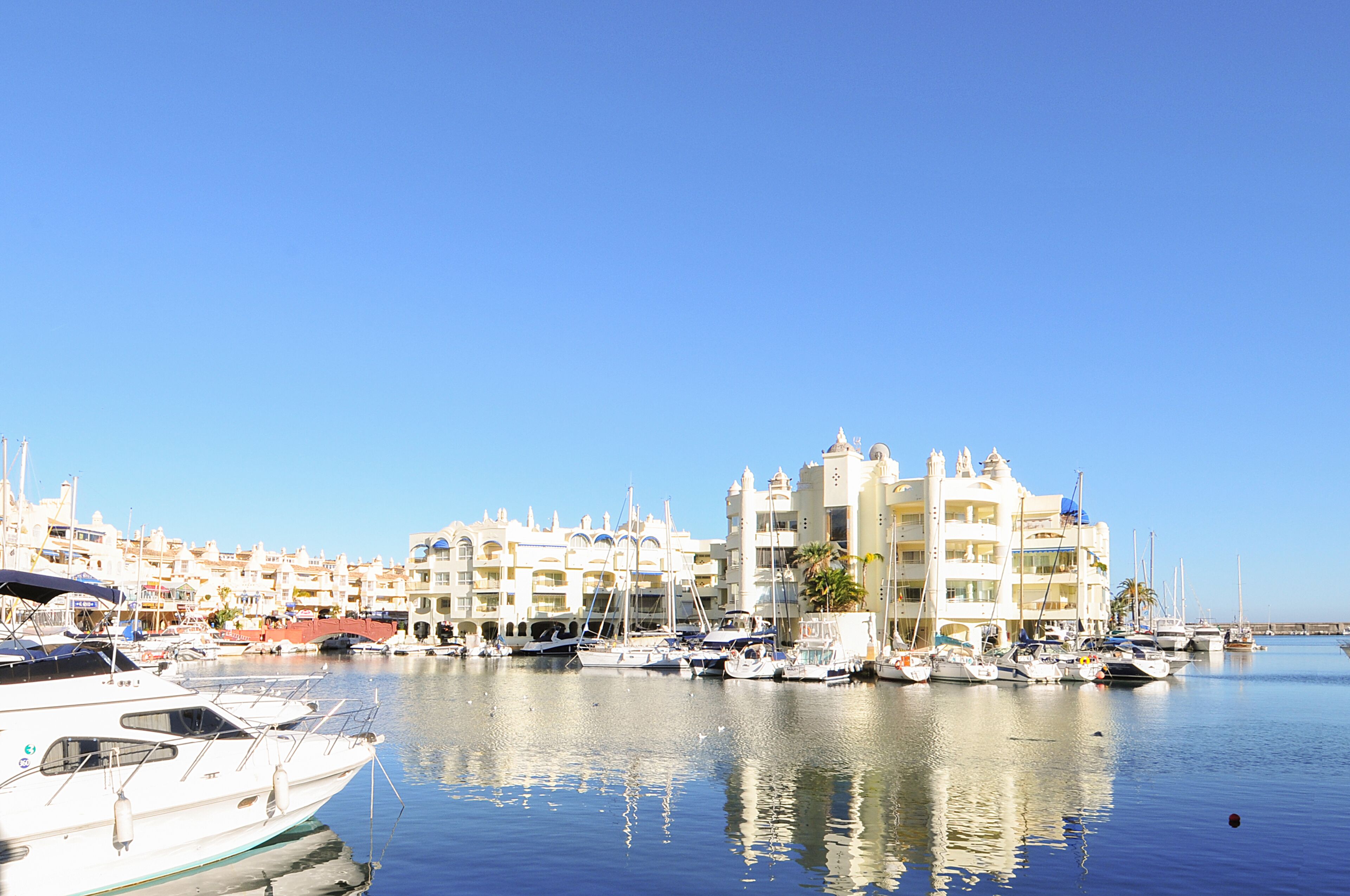 Un puerto deportivo con varias embarcaciones amarradas, un edificio grande con balcones y un cielo azul despejado.