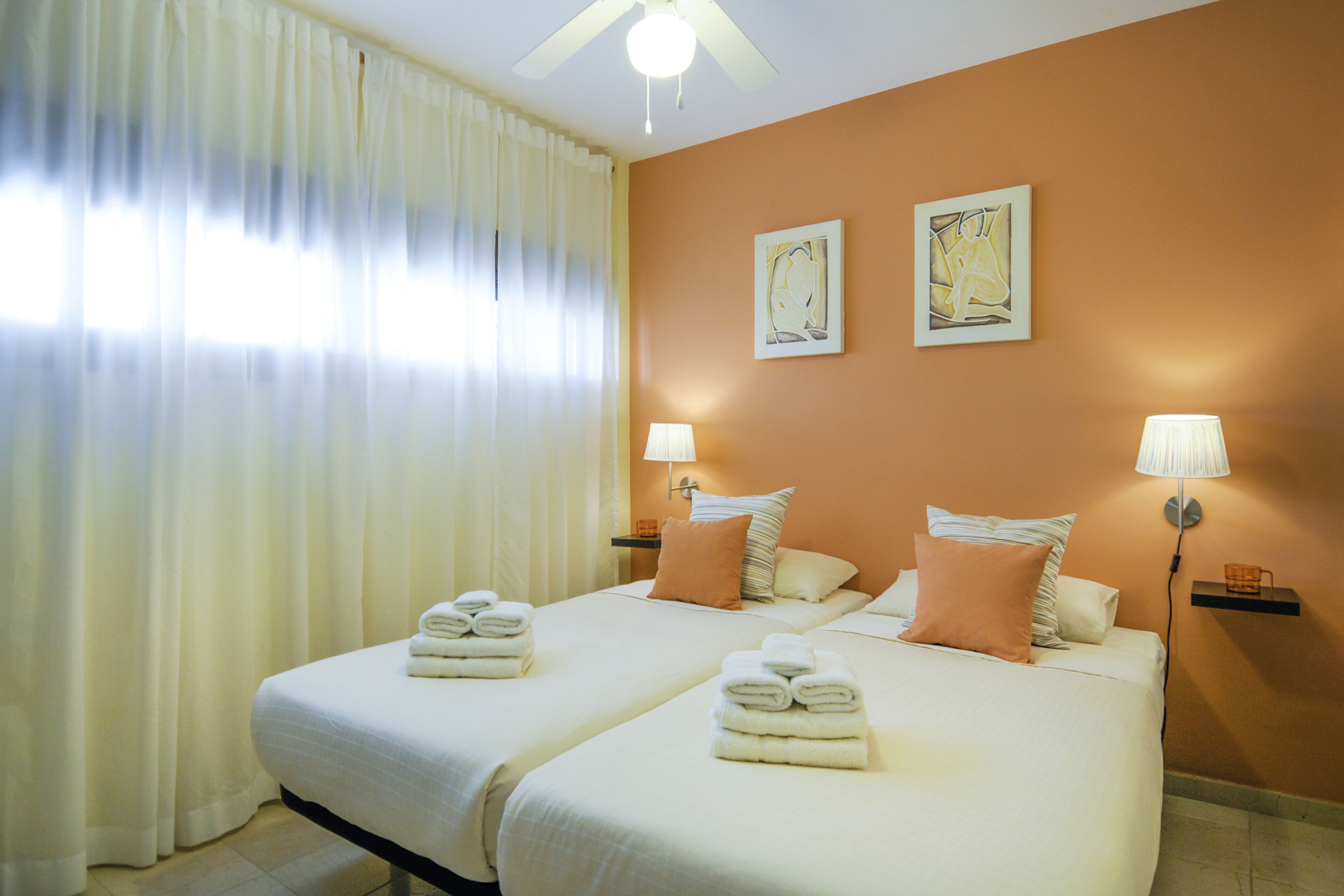 Camera d'albergo con due letti, lenzuola bianche, una ventola a soffitto, due lampade a parete e un quadro appeso al muro.