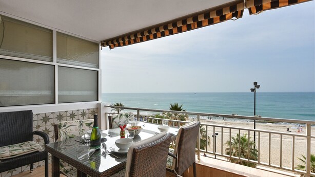A balcony with a table and chairs, overlooking a beach and the sea.