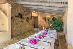 A long dining table set with white tablecloths, plates, and glasses, in a rustic stone-walled room with a green wheelbarrow in the background.