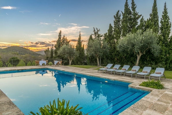 A swimming pool with lounge chairs and tables, surrounded by trees and a view of hills.