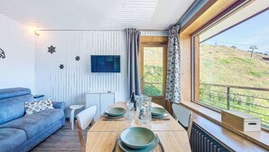 A cozy living room with a wooden dining table set for four, a blue sofa, and a TV mounted on the wall. - Horizon 2000 (Le Lac) by Interhome (Tignes)