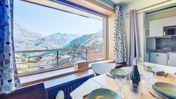 A dining area with a table set for two, a view of mountains and a village through large windows, and a kitchen area visible to the right. - Horizon 2000 (Le Lac) by Interhome (Tignes)