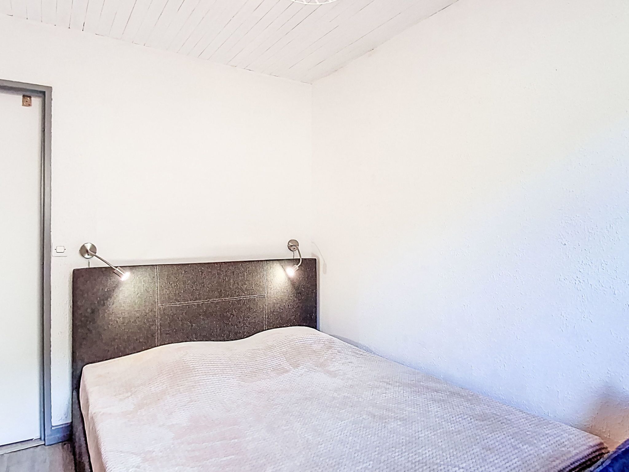 A single bed with a dark headboard and a light-colored mattress in a room with white walls and a wooden ceiling.