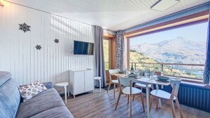 A cozy room with a sofa, a dining table with chairs, a TV mounted on the wall, and a large window offering a mountain view. - Horizon 2000 (Le Lac) by Interhome (Tignes)