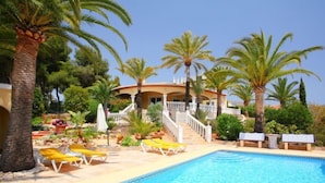 A pool area with lounge chairs, palm trees, and a building with a staircase leading to a balcony. - Bombilla by Interhome (Moraira)
