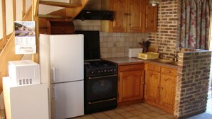 A compact kitchen with a refrigerator, stove, oven, wooden cabinets, and a brick wall.