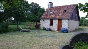 Una pequeña casa de piedra con techo inclinado y chimenea, rodeada por un césped con una silla y un parterre de flores.