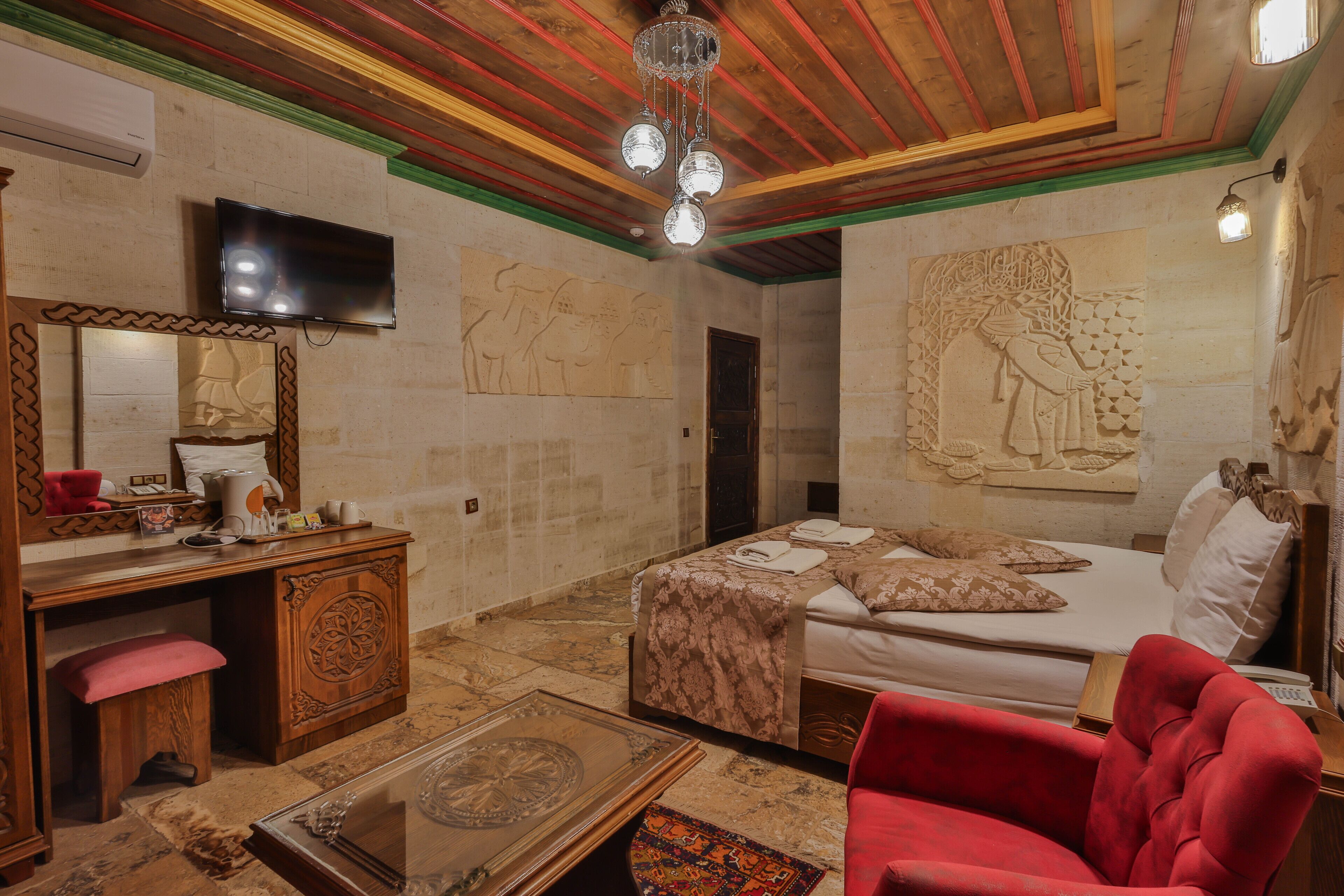 Habitación de hotel con techo de madera, televisor de pantalla plana, cama con colcha estampada, sillón rojo y un espejo decorativo.
