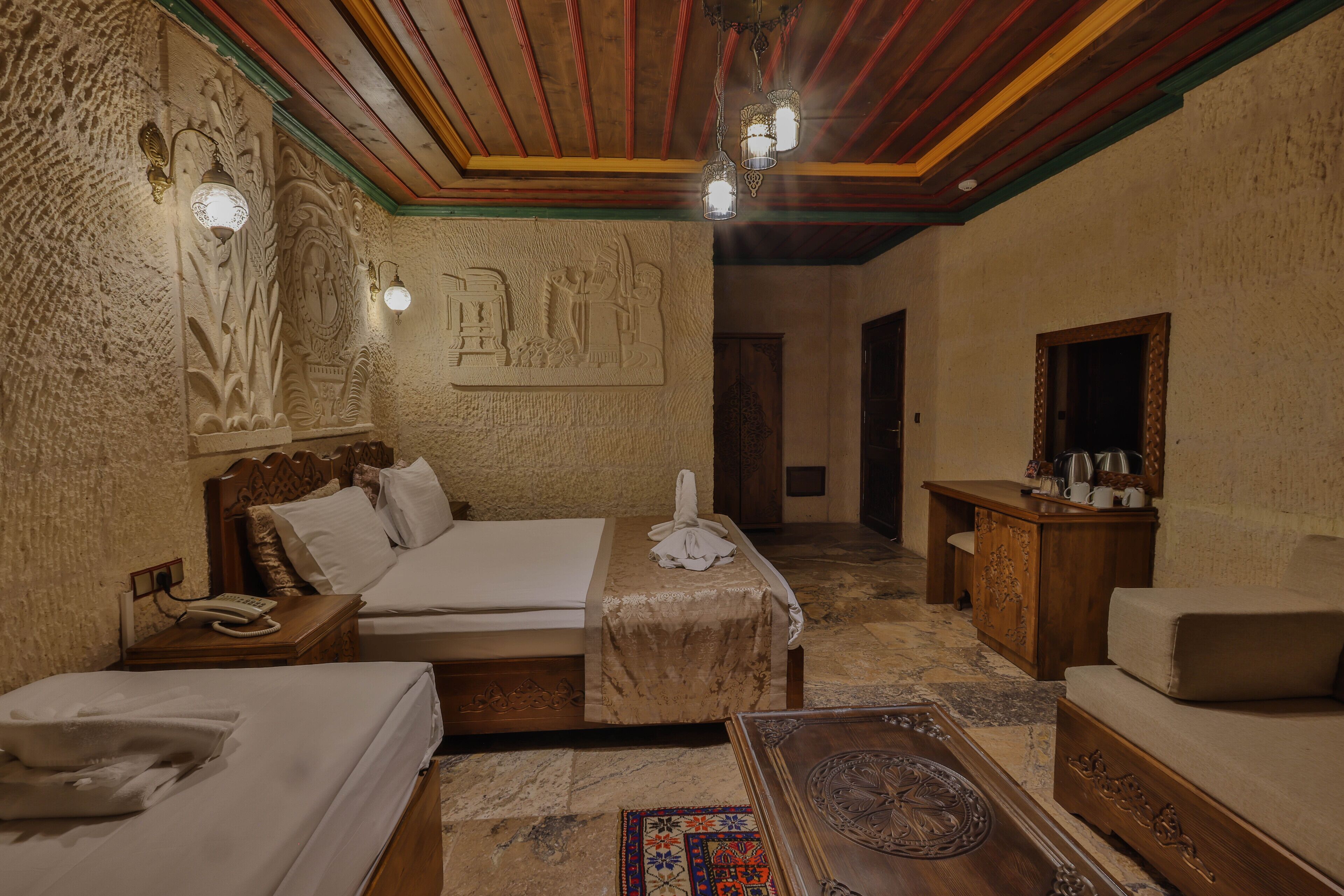 A hotel room with a wooden bed, a wooden nightstand with a telephone, a small table with a lamp, and a decorative wall carving.