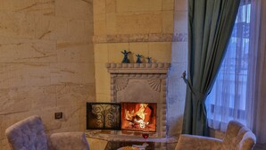 A cozy room with a fireplace, two armchairs, a table with a plate and a glass, and a patterned rug. - Feel Cappadocia Stone House (Nevsehir)