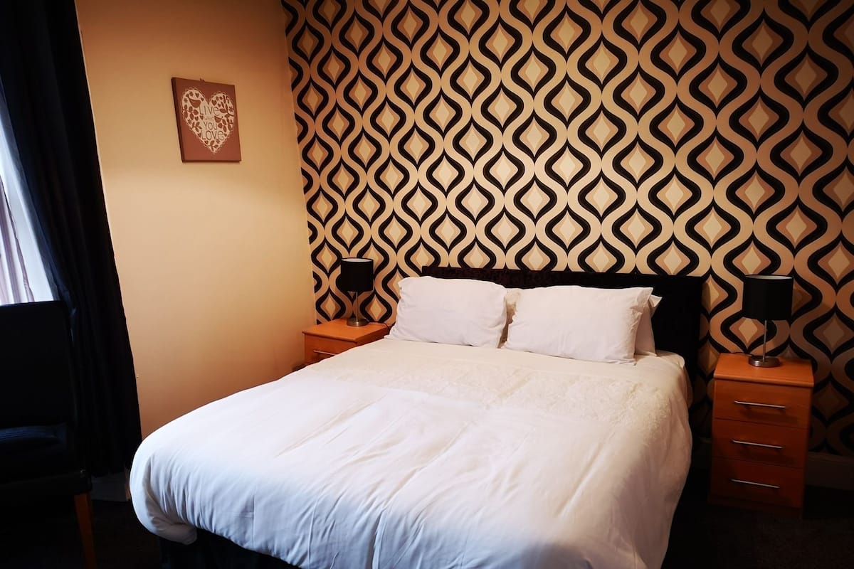 A hotel room with a patterned wallpaper, a bed with white bedding, two bedside tables with lamps, and a small artwork on the wall.