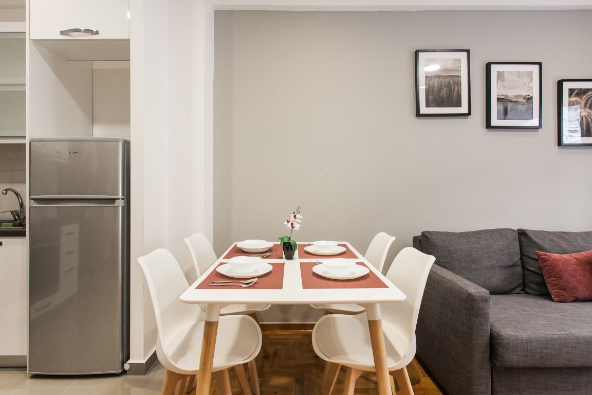 A modern kitchen with a stainless steel refrigerator, a dining table with white chairs, and a grey sofa with a red pillow.