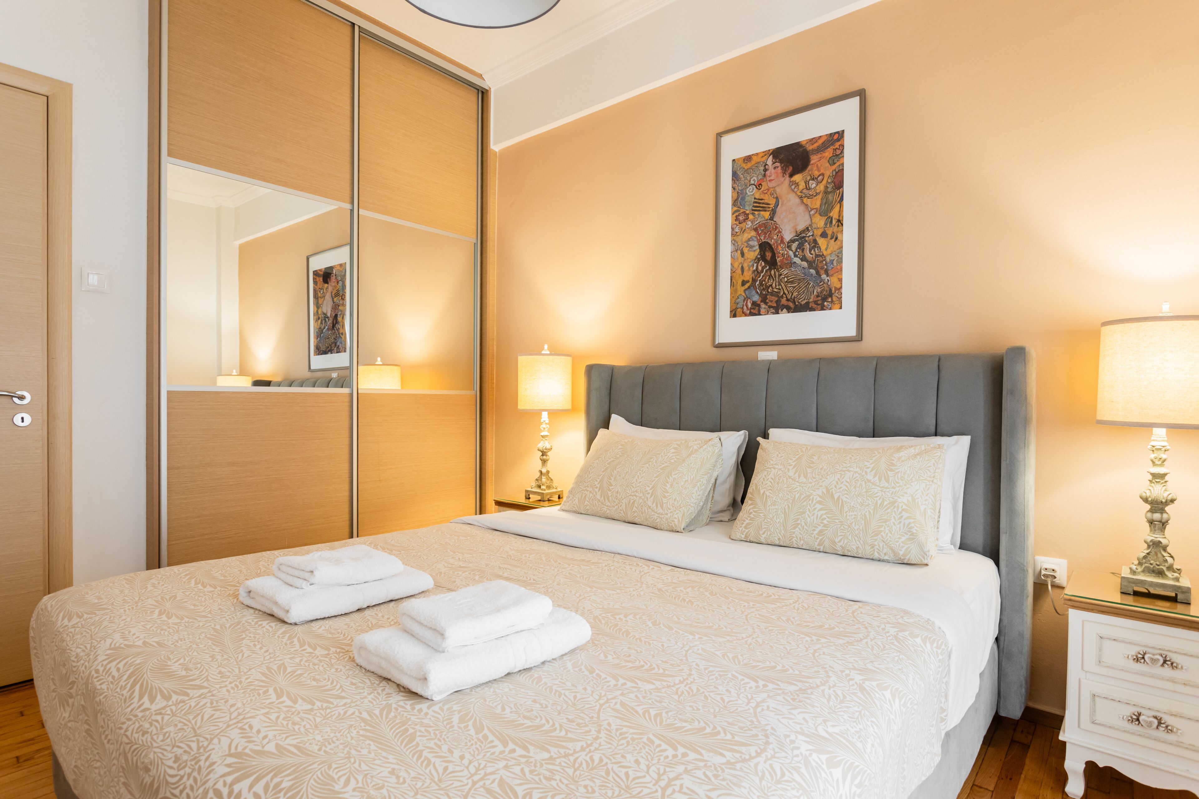A neatly made bed with white linens and folded towels, a grey upholstered headboard, a bedside table with a lamp, and a framed artwork above the bed.