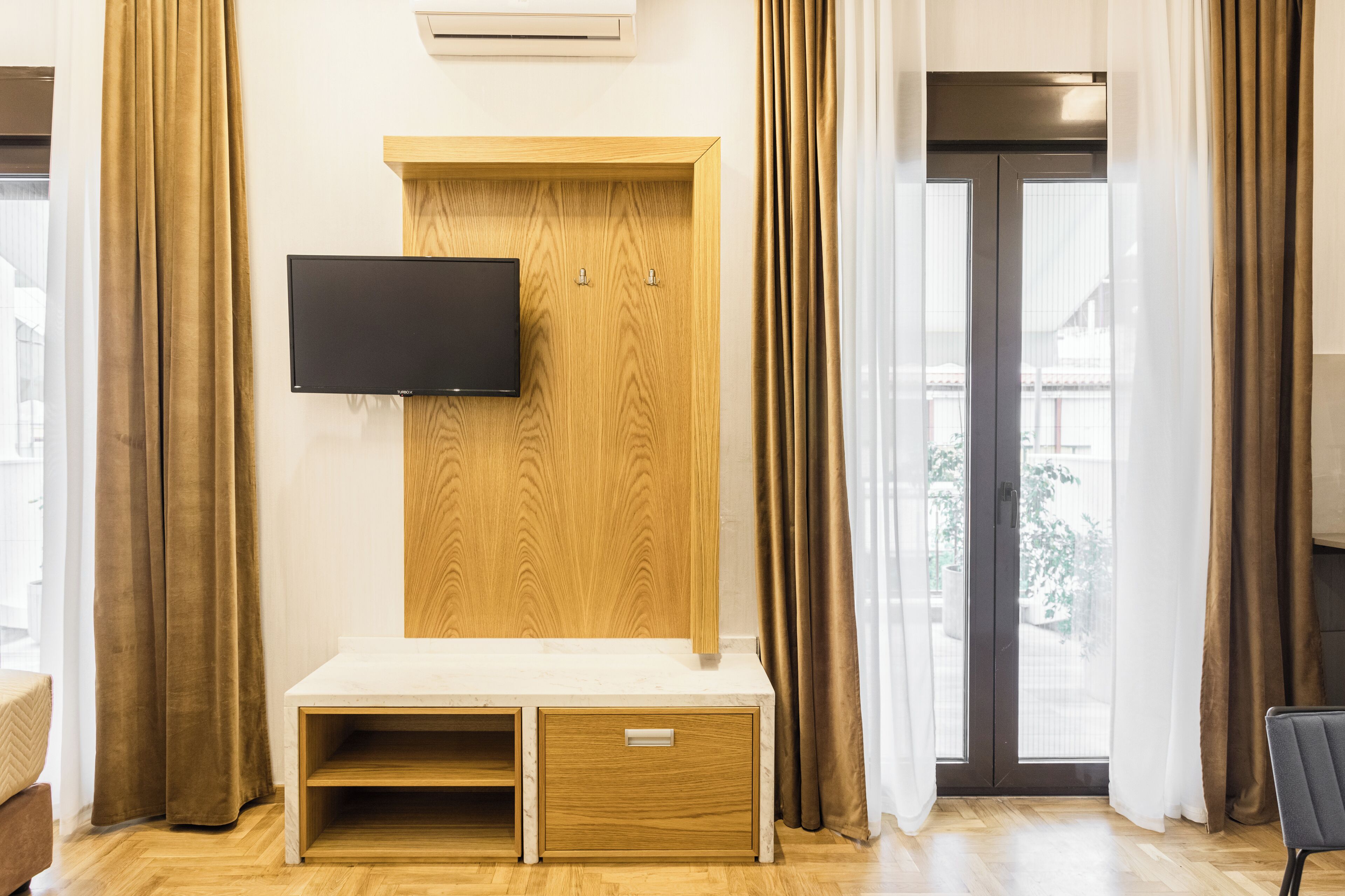 A modern hotel room with a wooden TV stand, a flat-screen TV, a small seating area, and a sliding glass door to the outside.