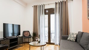 A living room with a flat-screen TV, a sofa, a coffee table, and a balcony with curtains. - Nicholas Point Apartments and Rooms (Athens)