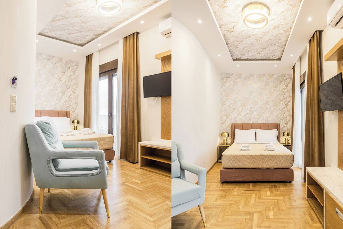 Habitación de hotel moderna con suelo de madera, cama con cabecera, zona de estar con una silla, televisor de pantalla plana montado en la pared e iluminación empotrada en el techo.