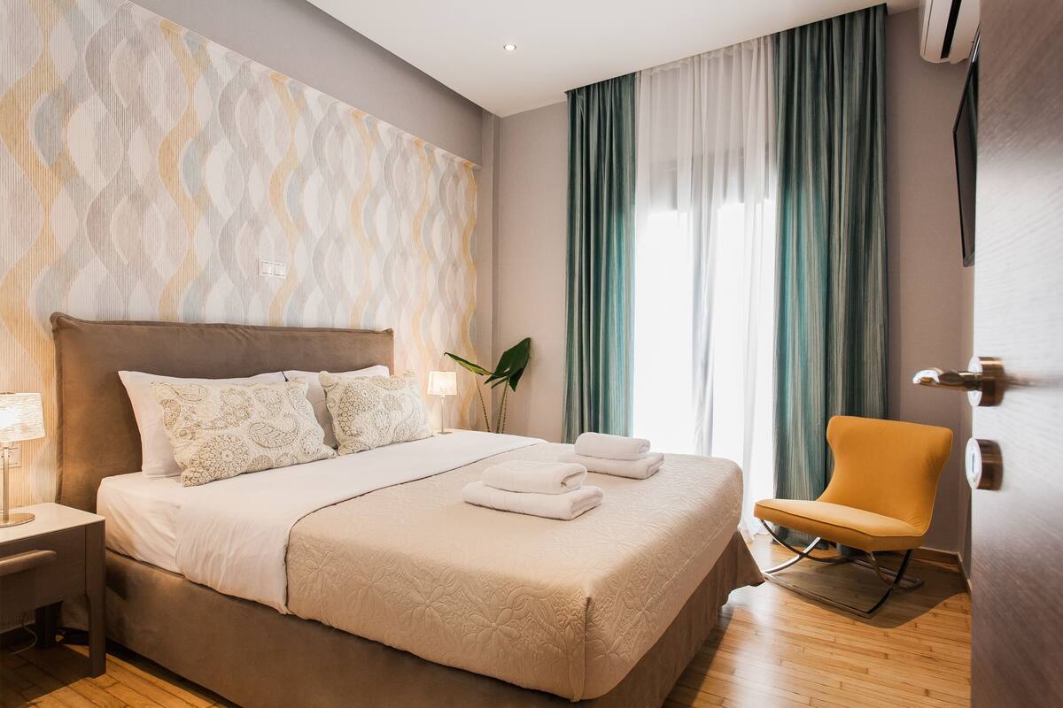 Una cama bien hecha con una colcha color beige y almohadas, una mesita de noche con una lámpara, una silla y una ventana grande con cortinas.
