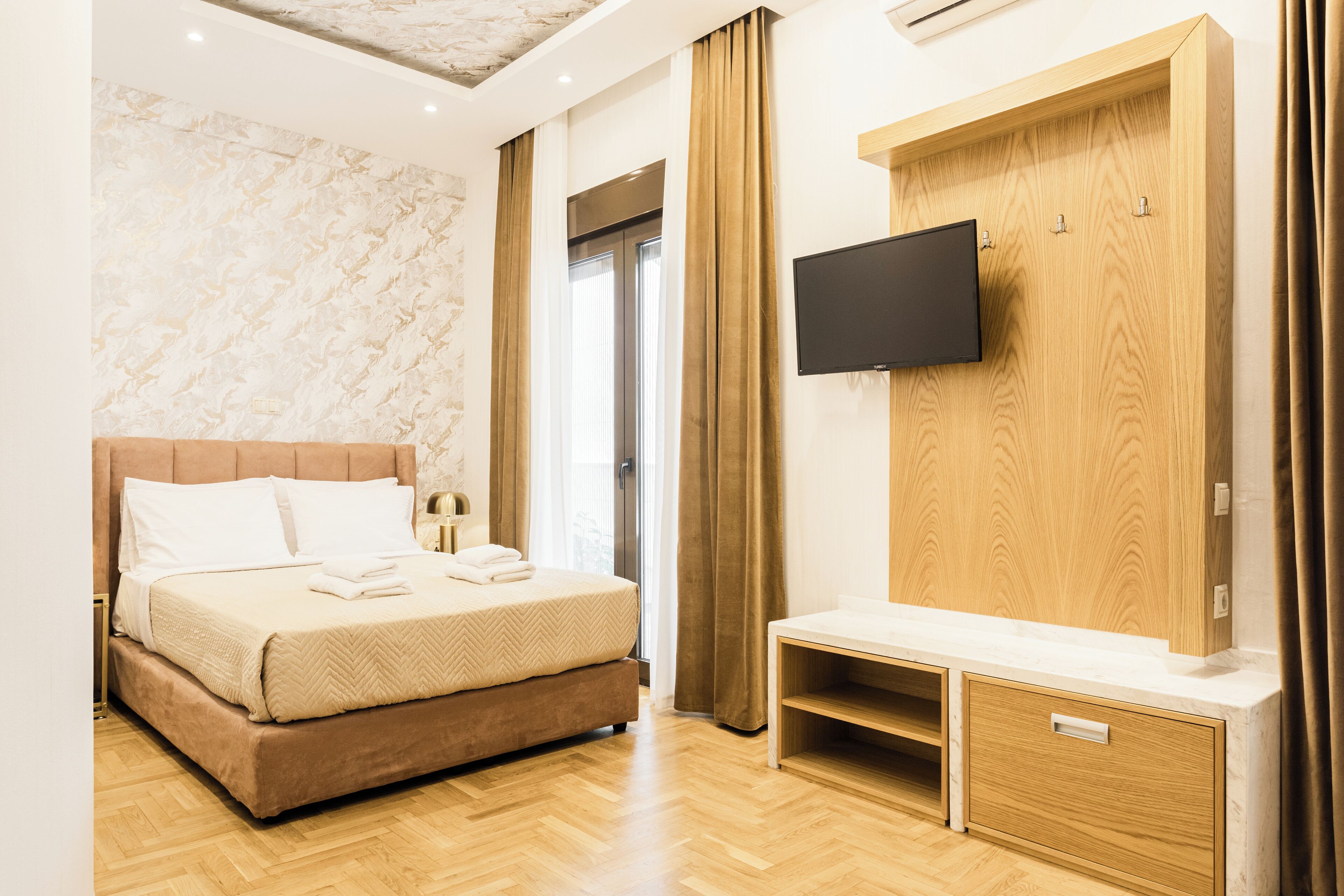 A modern hotel room with a large bed, a TV mounted on a wooden panel, and a wooden bench.