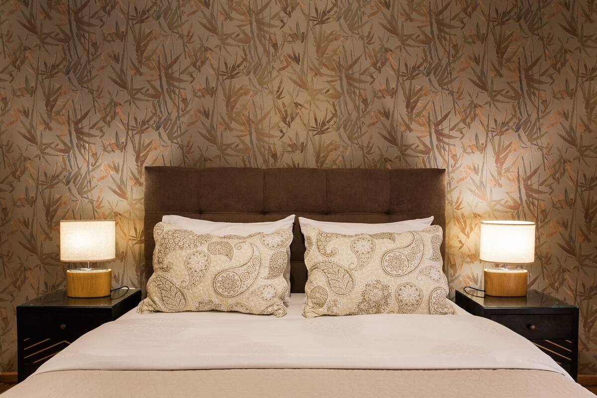 Una cama bien hecha con dos almohadones decorativos, un cabecero de madera y dos lámparas de noche sobre mesitas a juego.