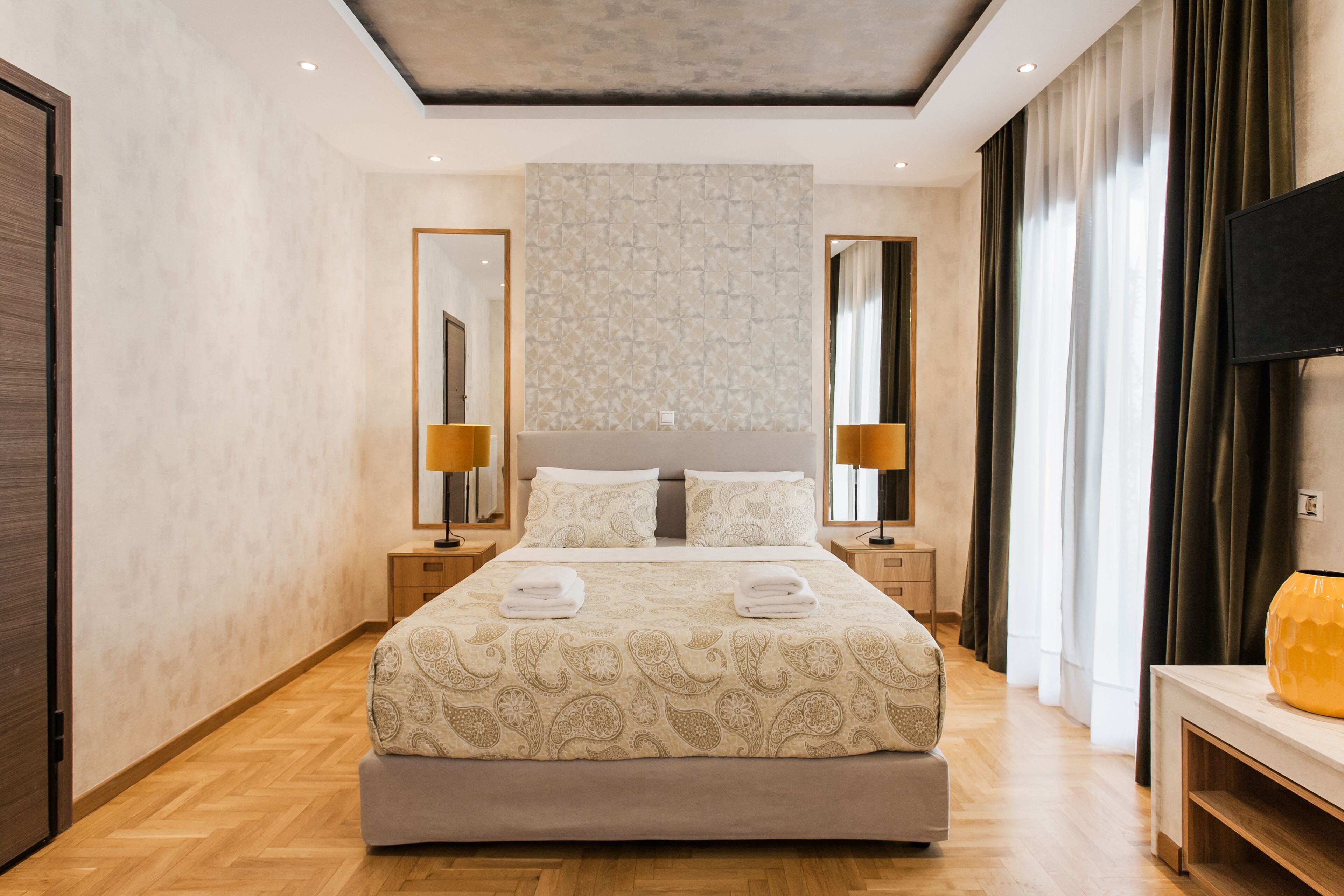A modern hotel room with a large bed, two bedside tables with lamps, a flat-screen TV, and a large window with curtains.