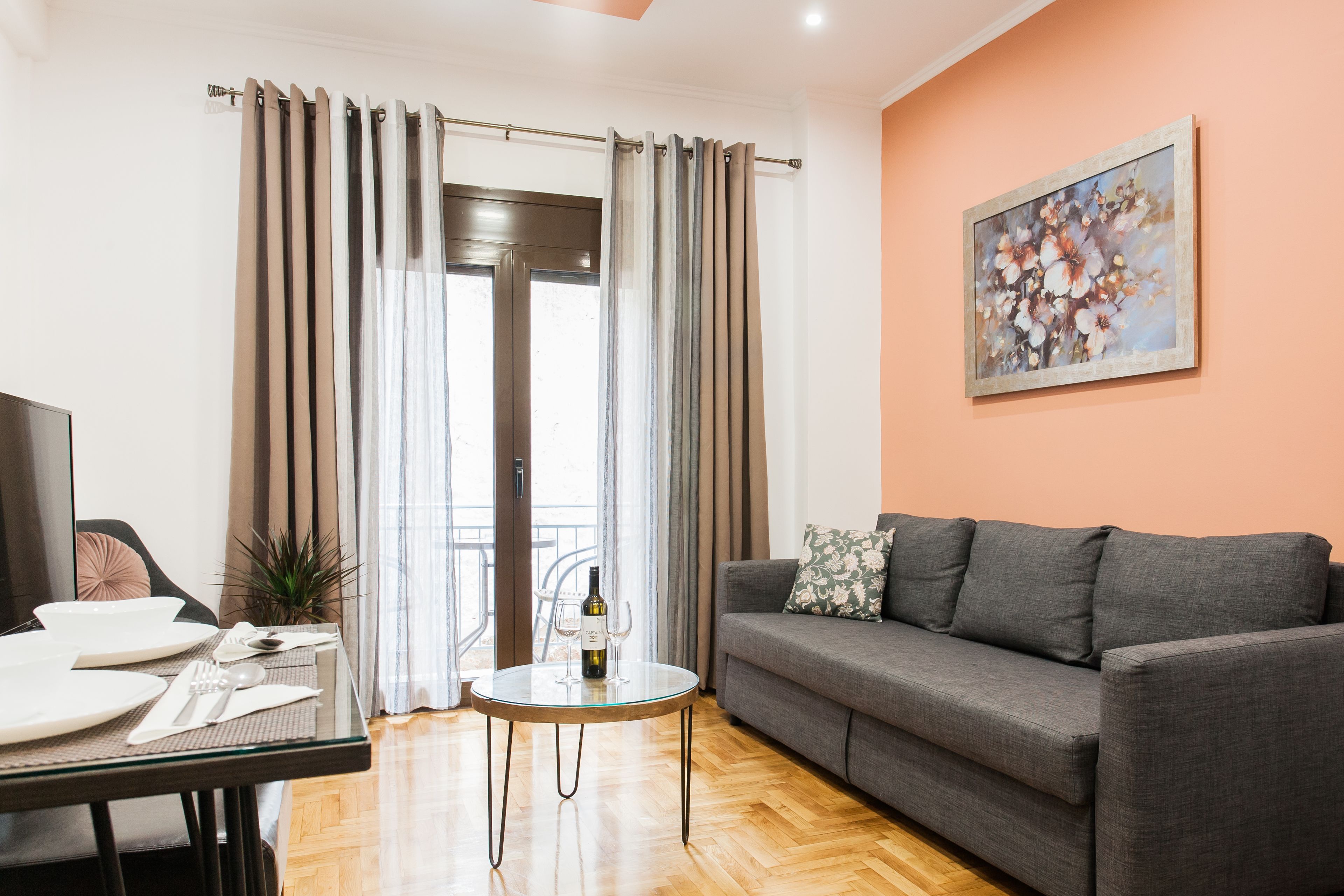 A living room with a grey sofa, a glass coffee table with a bottle and wine glass, a dining table set with plates and cutlery, and a painting on the wall.