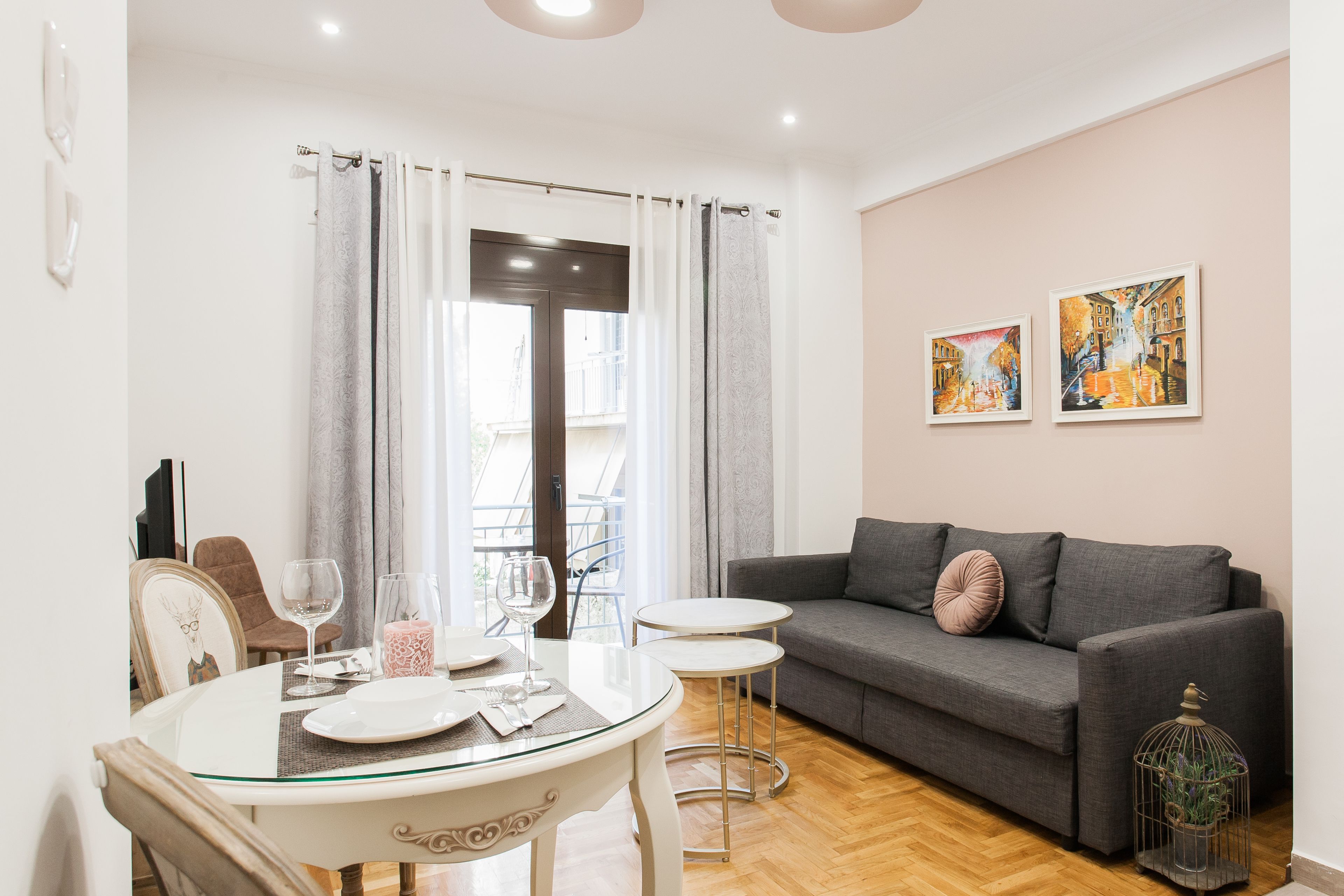 Una sala moderna con un sofá gris, una mesa de comedor de vidrio y dos mesitas auxiliares. Hay dos cuadros enmarcados en la pared y una jaula de pájaros en el suelo.