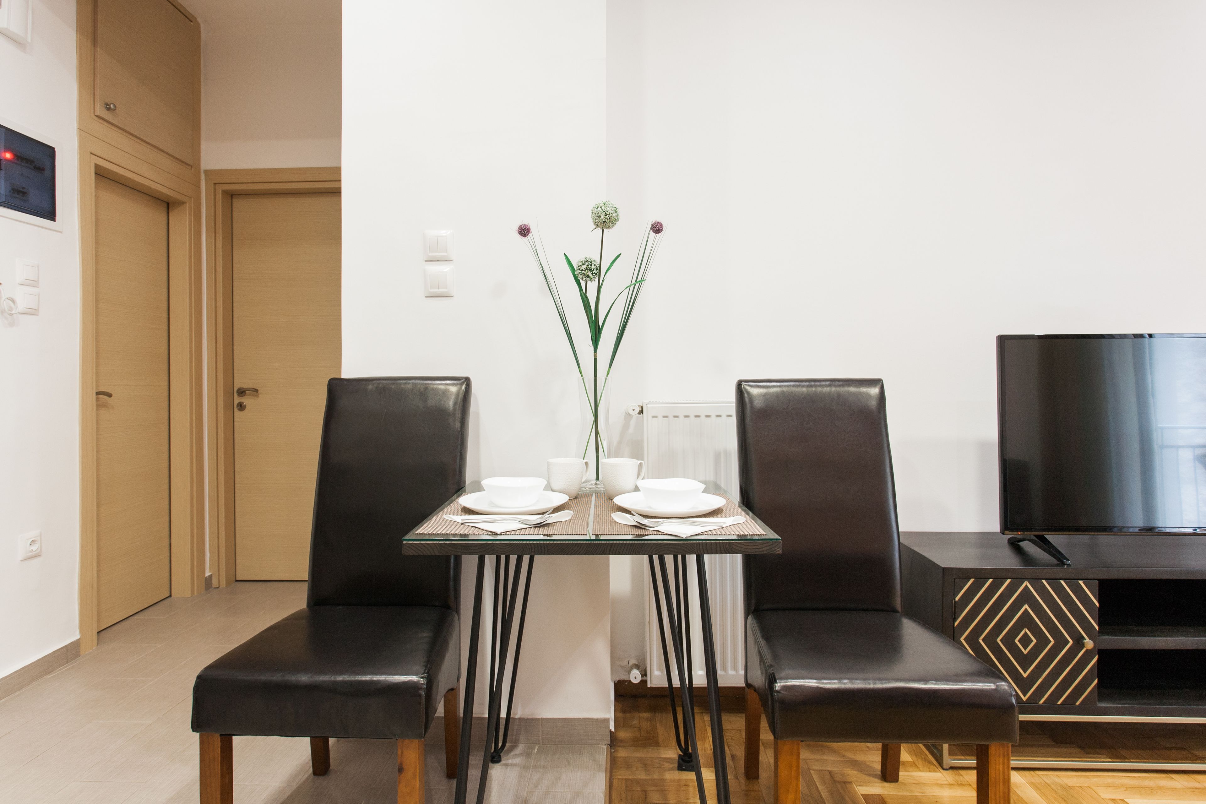 Un comedor moderno con una mesa de vidrio, dos sillas de cuero negro, un jarrón con flores y un televisor sobre un mueble negro.