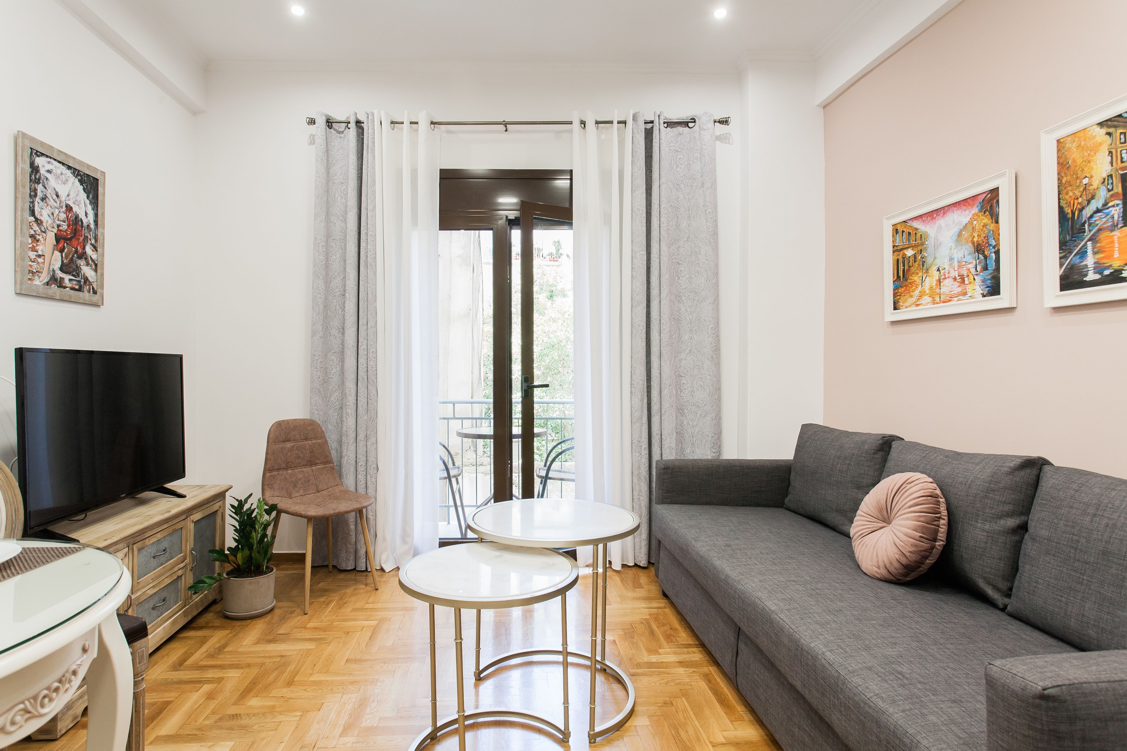 A modern living room with a grey sofa, a TV, a small table, and a chair. There are two paintings on the wall and a balcony with curtains.