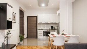 A modern kitchen with white cabinets, a dining area with white chairs, and a grey sofa. - Nicholas Point Apartments and Rooms (Athens)