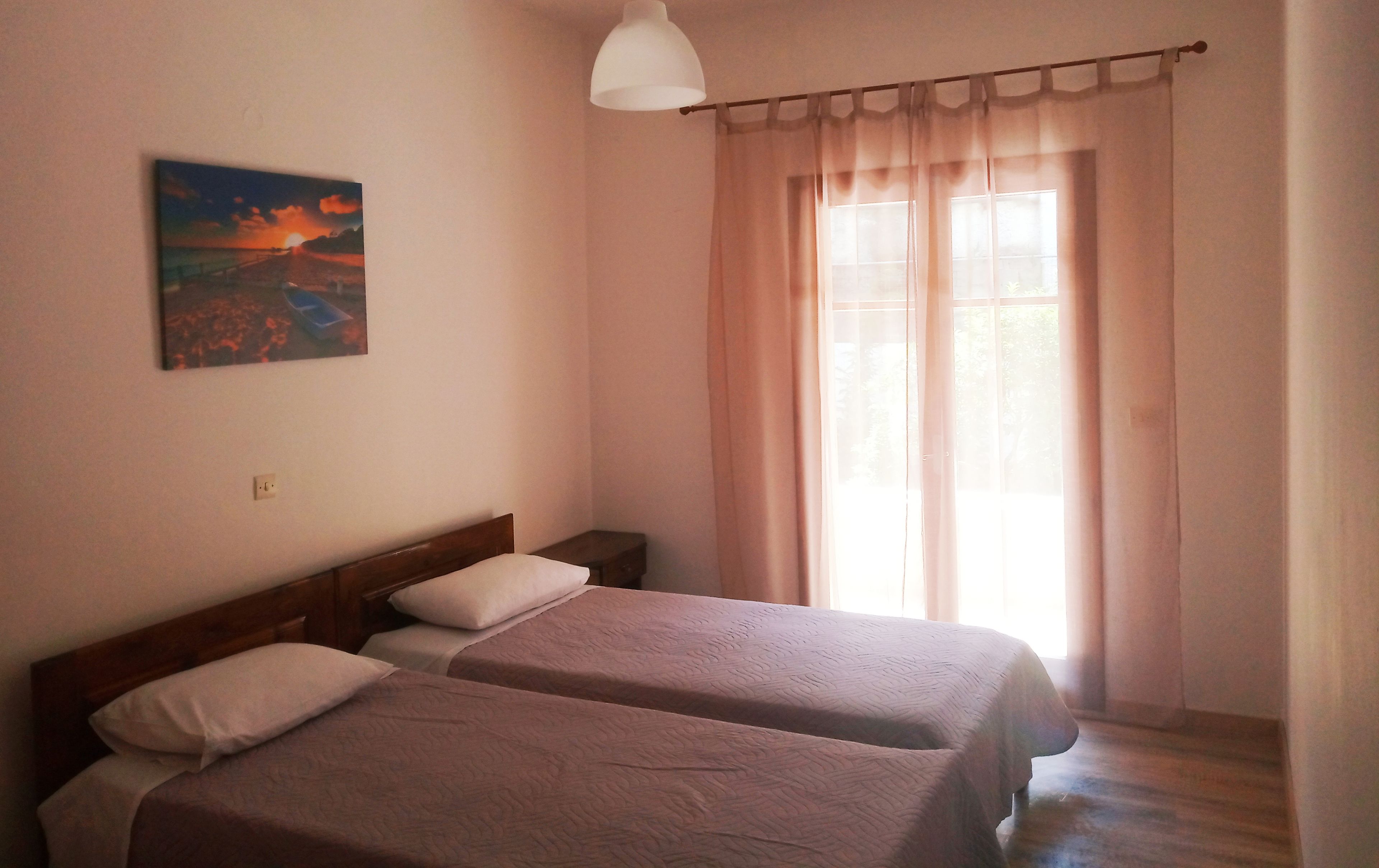 A room with two beds, a painting on the wall, a window with curtains, and a hanging light fixture.