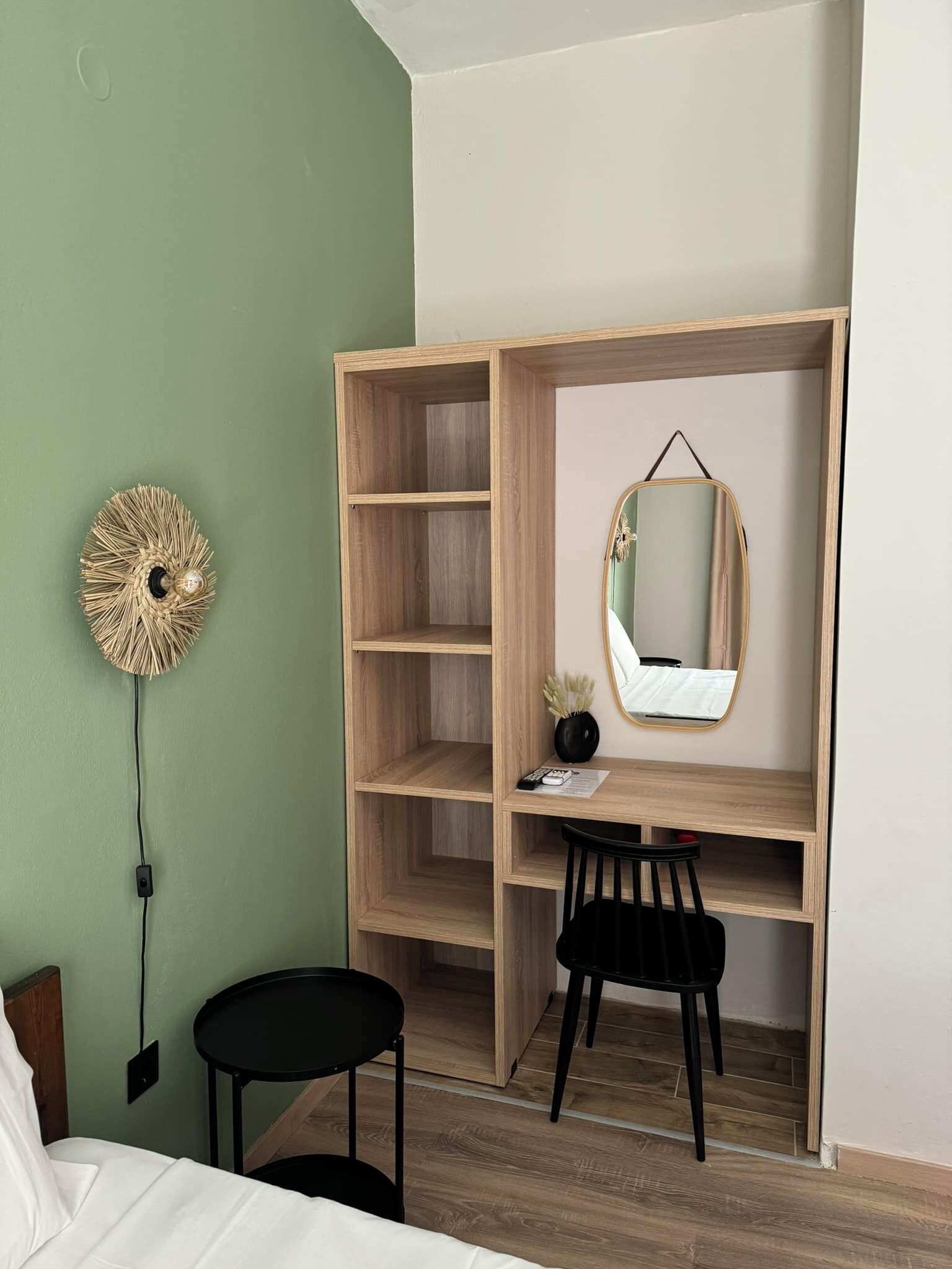 A bedroom with a wooden wardrobe, a small desk, a black chair, a round mirror, and a wall-mounted light fixture.