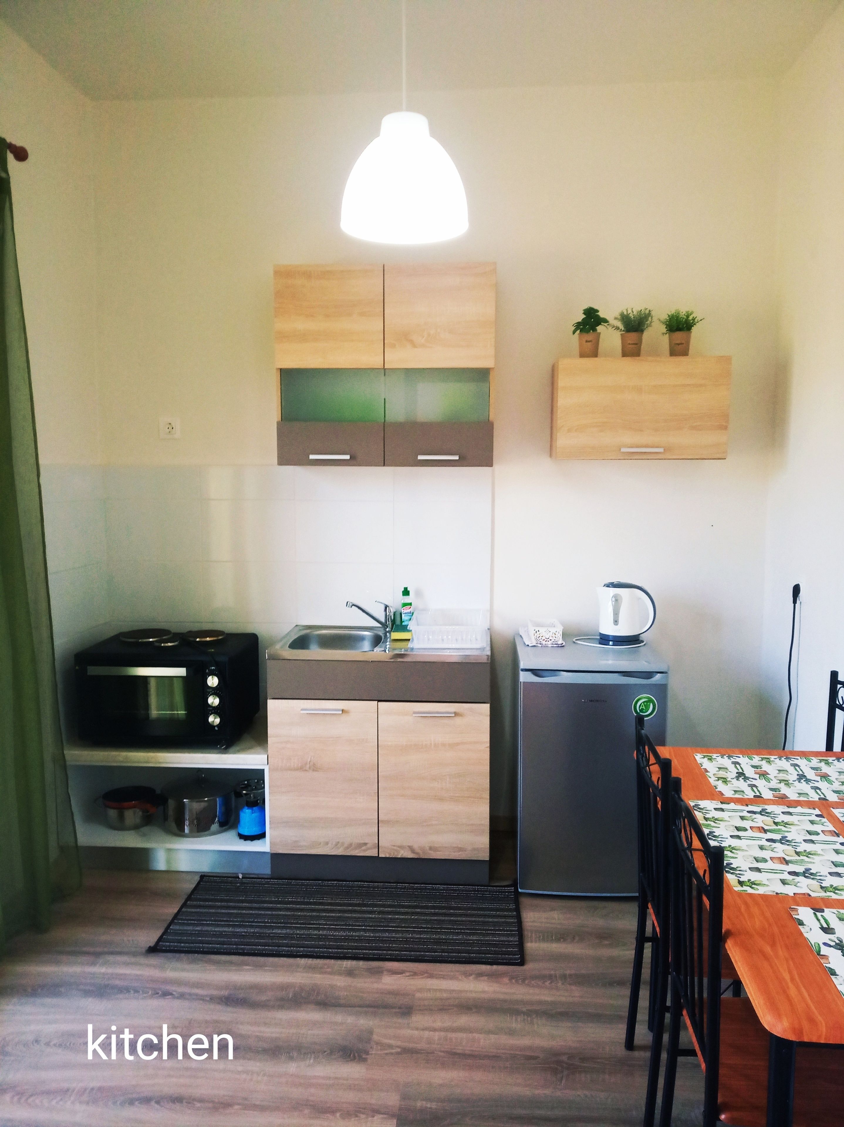 A compact kitchen with wooden cabinets, a microwave, a kettle, and a small refrigerator.