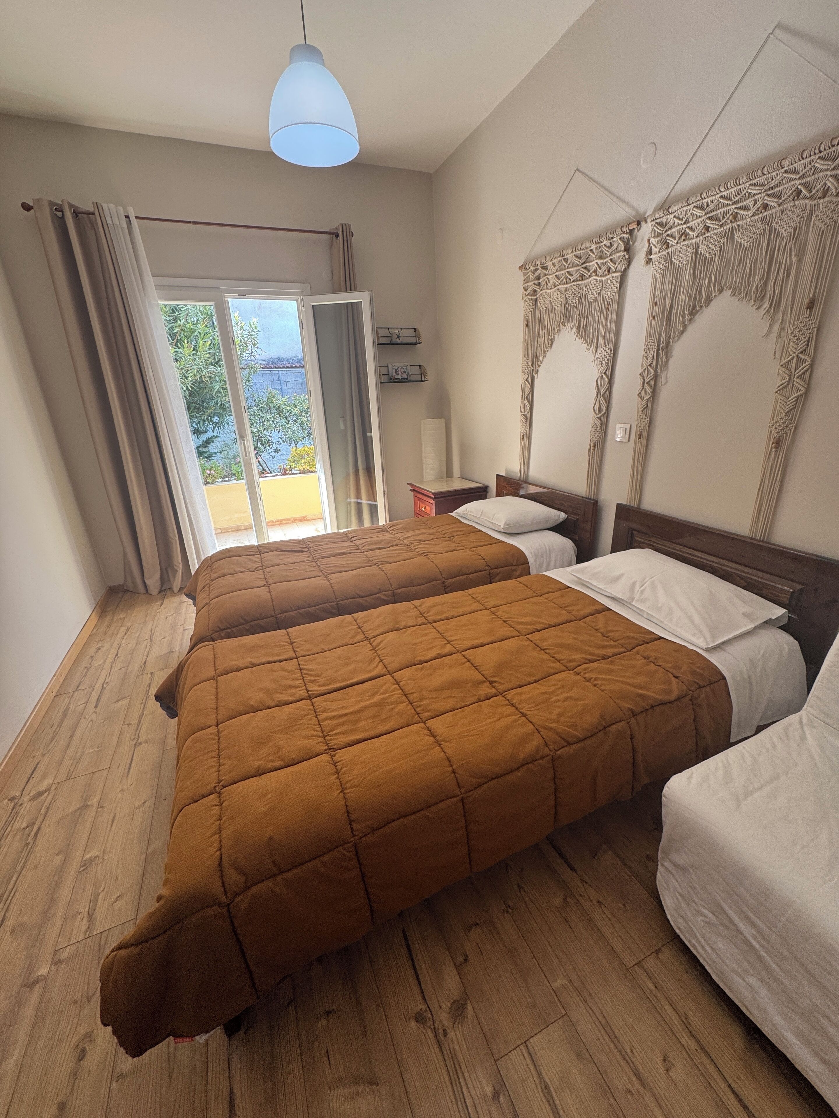 Une chambre d’hôtel avec deux lits, un plancher en bois, un luminaire au plafond, une fenêtre avec des rideaux et une vue sur la verdure.