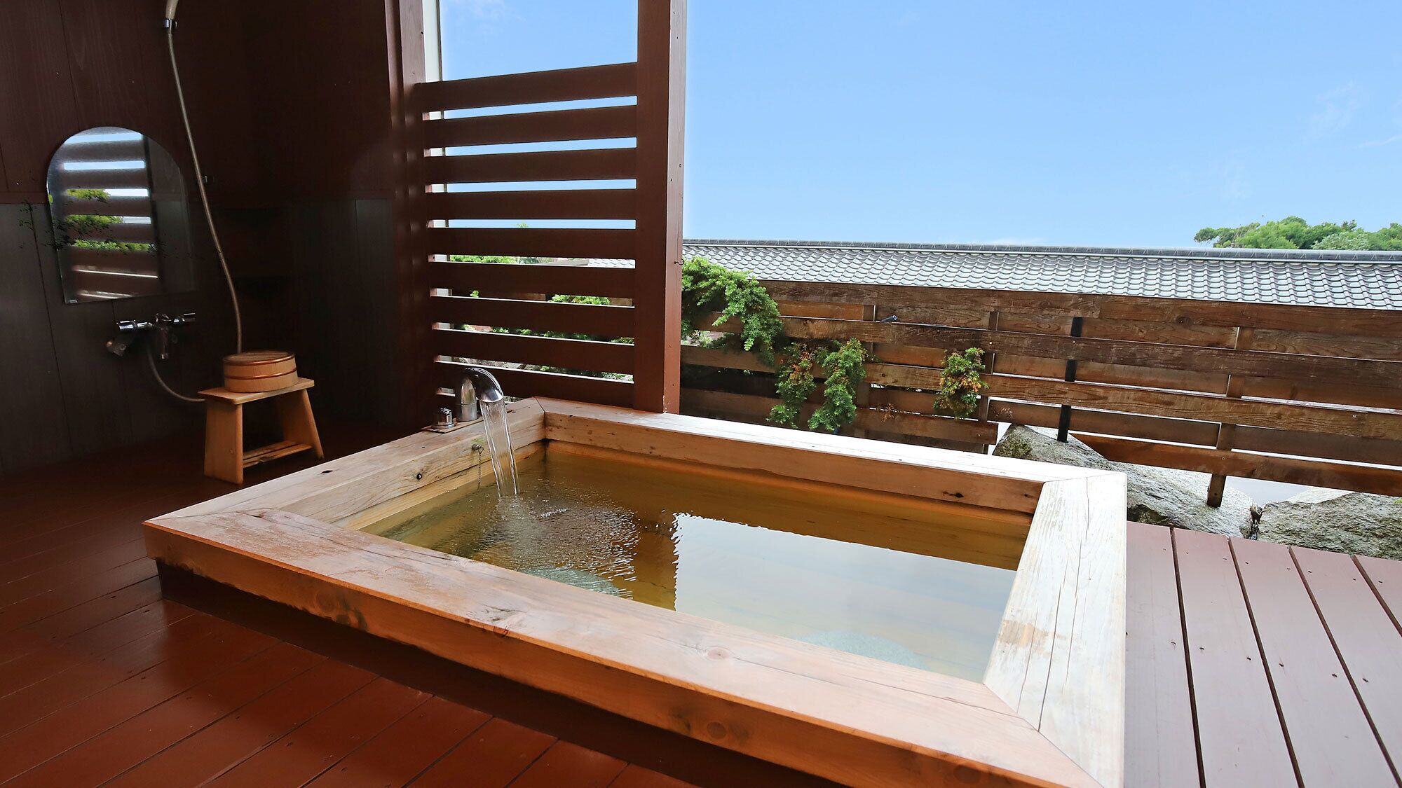 Una bañera de hidromasaje de madera con vista a un tejado tradicional con tejas y follaje.