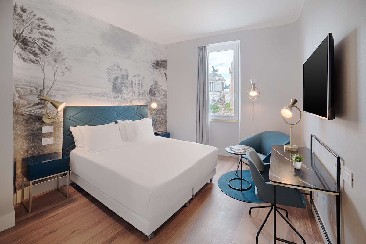A modern hotel room with a bed, a desk, a chair, a TV, and a mural on the wall.