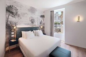 A hotel room with a large bed, a nightstand, a wall-mounted lamp, and a view of the city through a window.