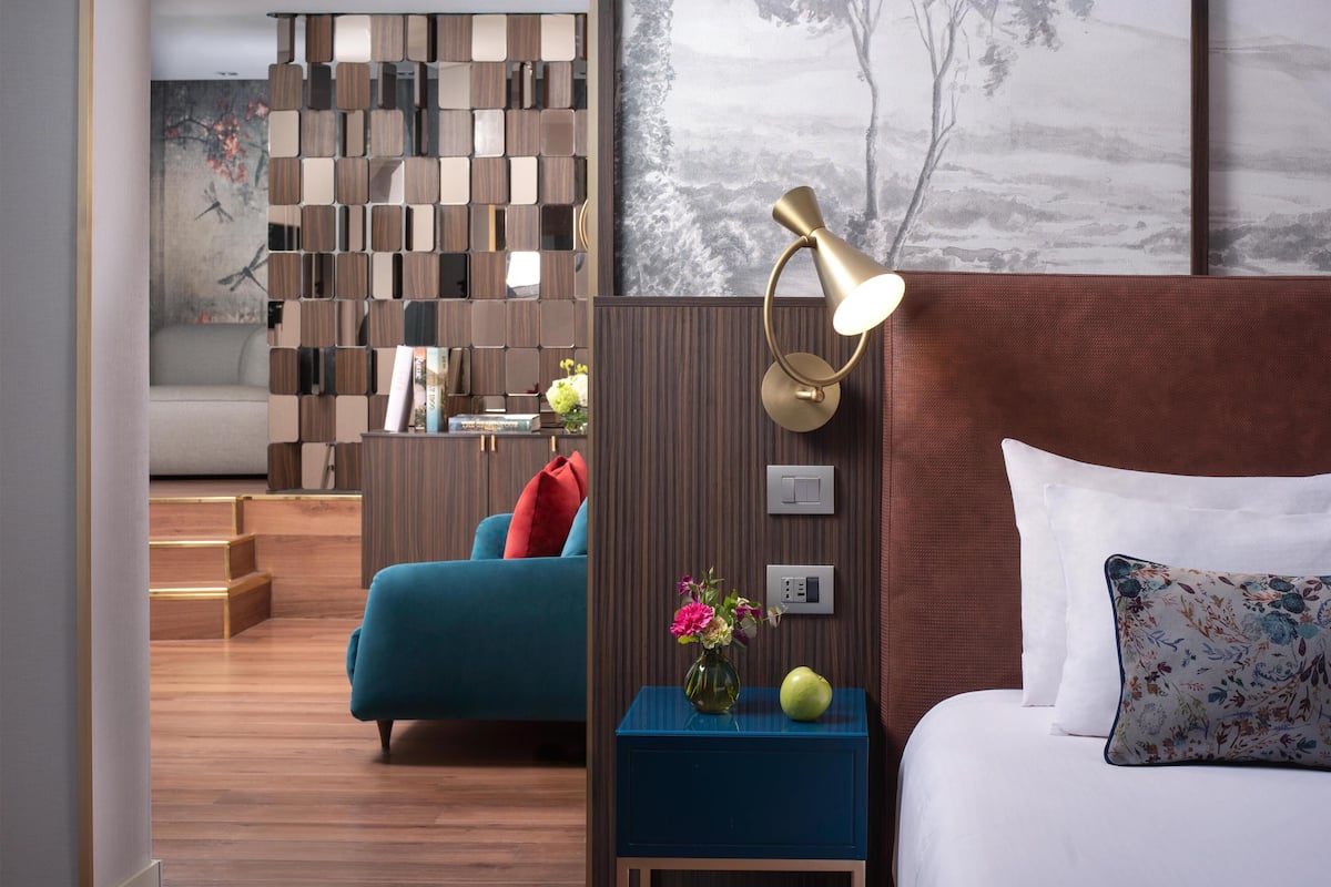 A modern hotel room with a wooden floor, a blue sofa, a nightstand with a lamp, and a bed with white bedding.