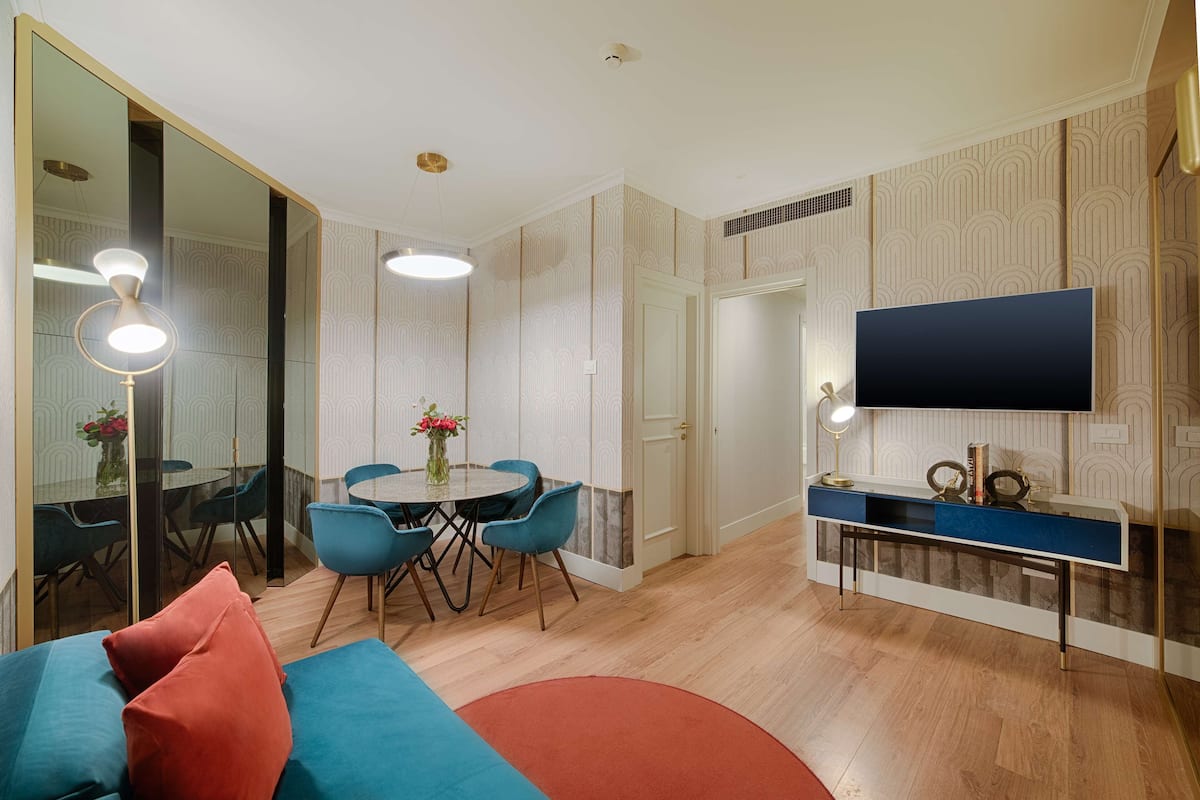 A modern living room with a round dining table, blue chairs, a sofa with red cushions, a TV on the wall, and a console table with decorative items.