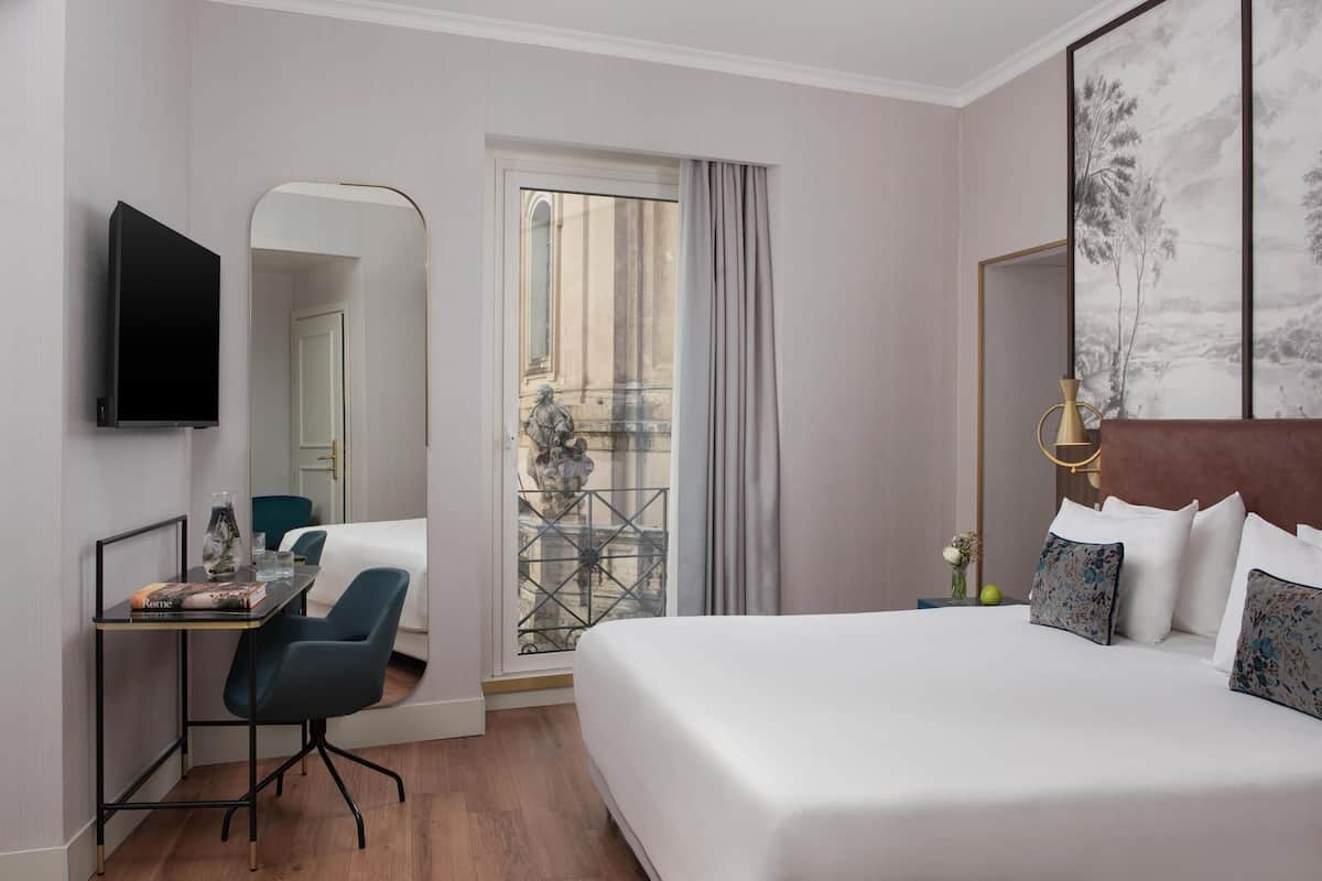 A hotel room with a bed, a desk with a chair, a TV, a window with a view of a building, and a painting on the wall.