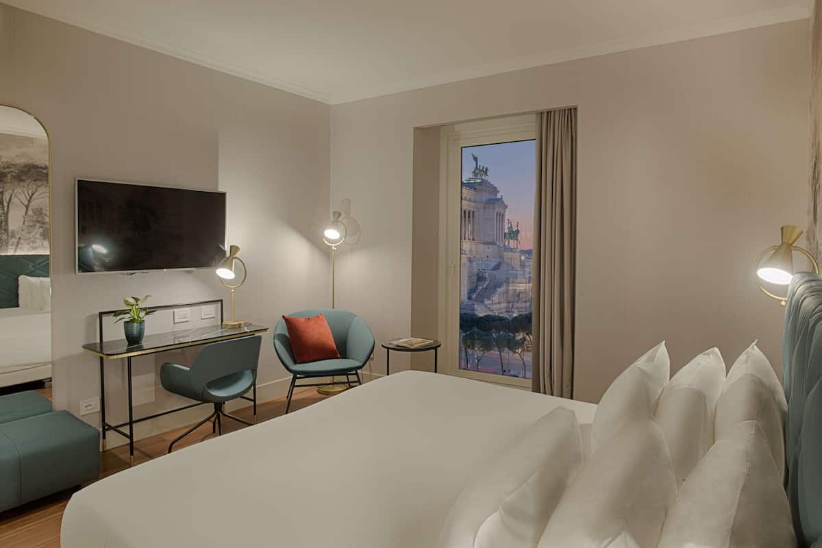 A hotel room with a bed, a desk, a chair, a TV, and a window with a view of a building.