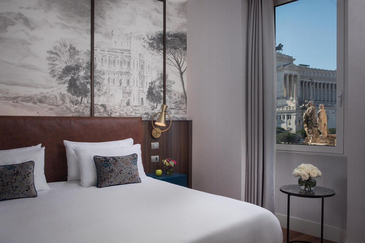 A hotel room with a large bed, a headboard, a nightstand with a vase of flowers, and a view of a building through a window.