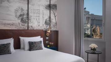 A hotel room with a large bed, a headboard, a nightstand with a vase of flowers, and a view of a building through a window.