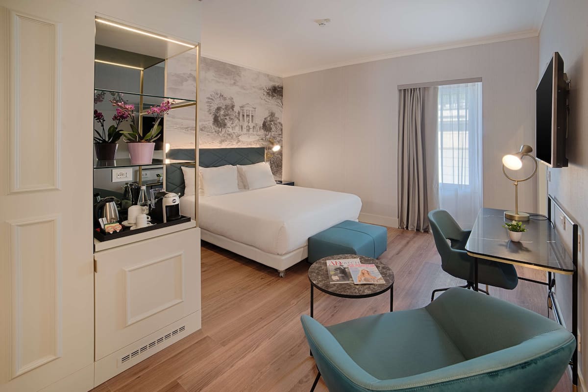 A hotel room with a bed, a desk, a chair, a small table, a mirror, a potted plant, and a wall with a mural.